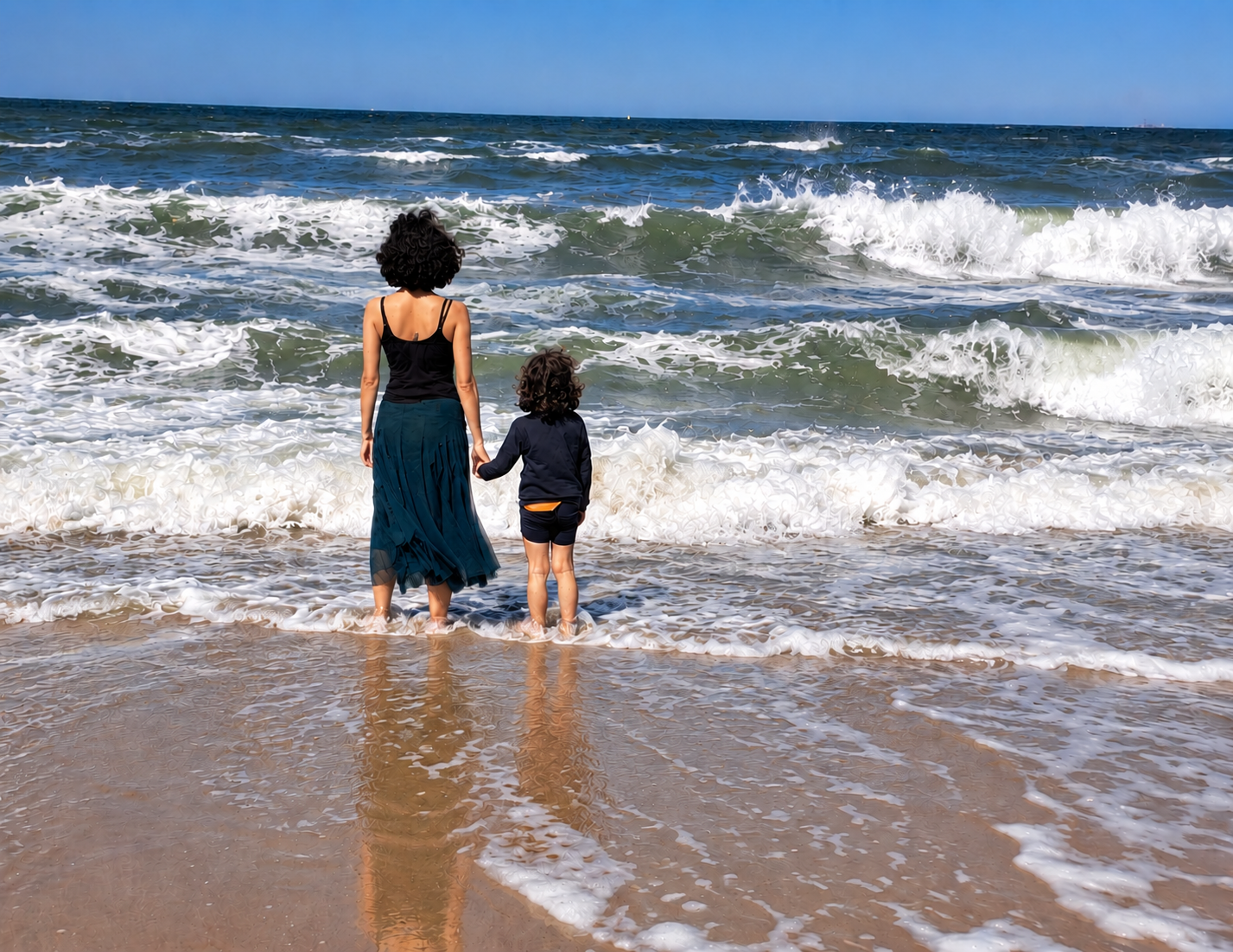 Kiran steht Hand in Hand mit ihrem Kind von hinten im Meer und schaut auf die Wellen